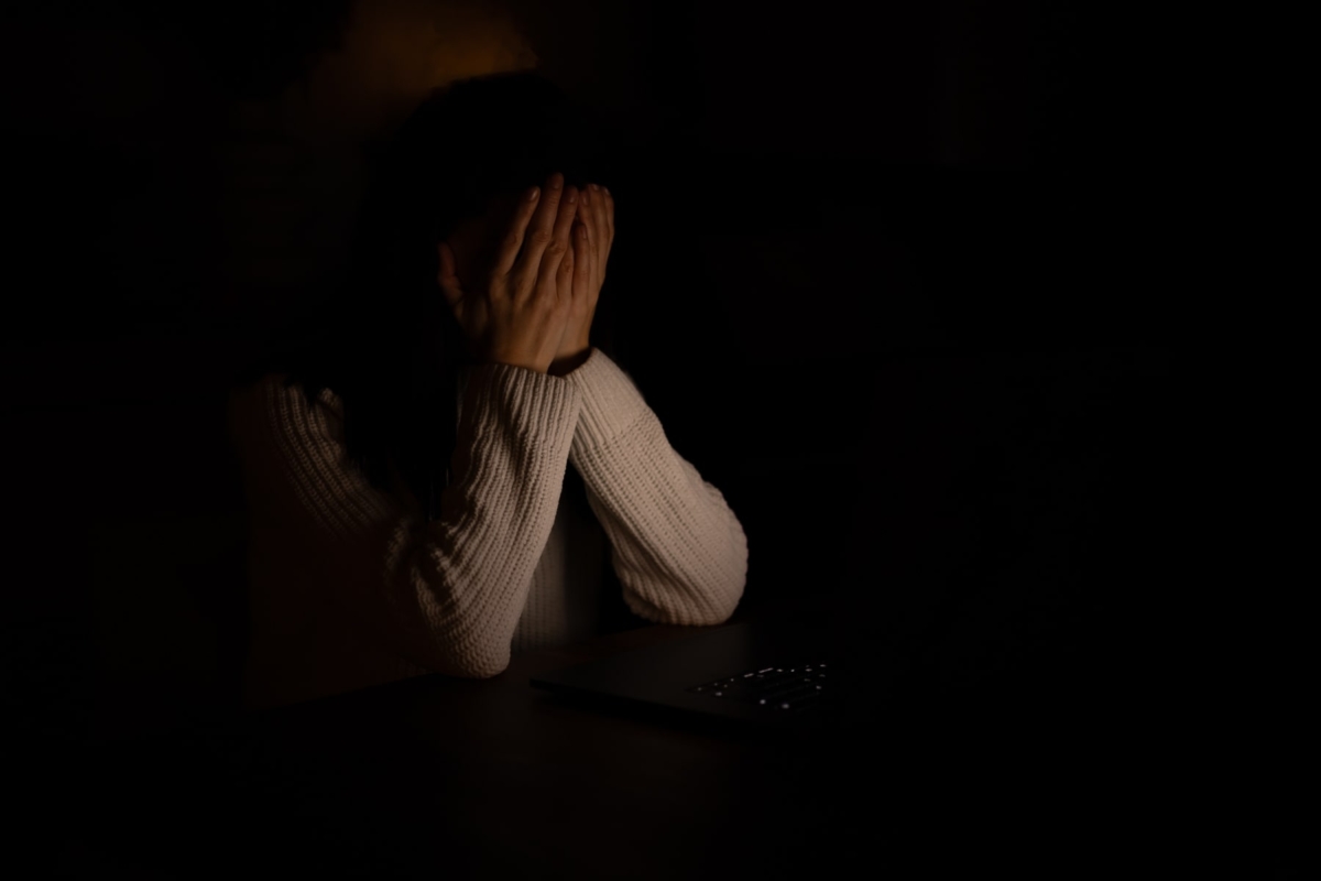 Kobieta zakrywa twarz dłońmi, siedząc przed laptopem w ciemnym pomieszczeniu