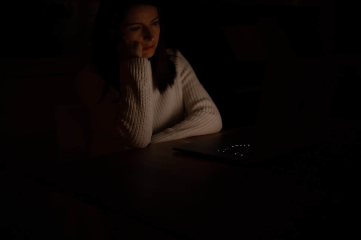 Kobieta w białym swetrze patrząca na laptopa, wspierając głowę na dłoni, w przygaszonym świetle.